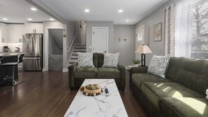 A modern living room with a sofa set, a coffee table, and a kitchen area with a refrigerator. - 5 Bedrooms w. TVs in every room & Garage Parking (Overland Park)