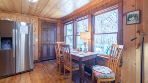 A wooden cabin interior with a stainless steel refrigerator, wooden dining table with chairs, and a window overlooking a snowy landscape. - Charming 2-bedroom cottage near Highlands and attractions (Scaly Mountain)