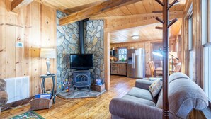 A wooden interior with a stone fireplace, a television, a sofa, and a kitchen area. - Charming 2-bedroom cottage near Highlands and attractions (Scaly Mountain)