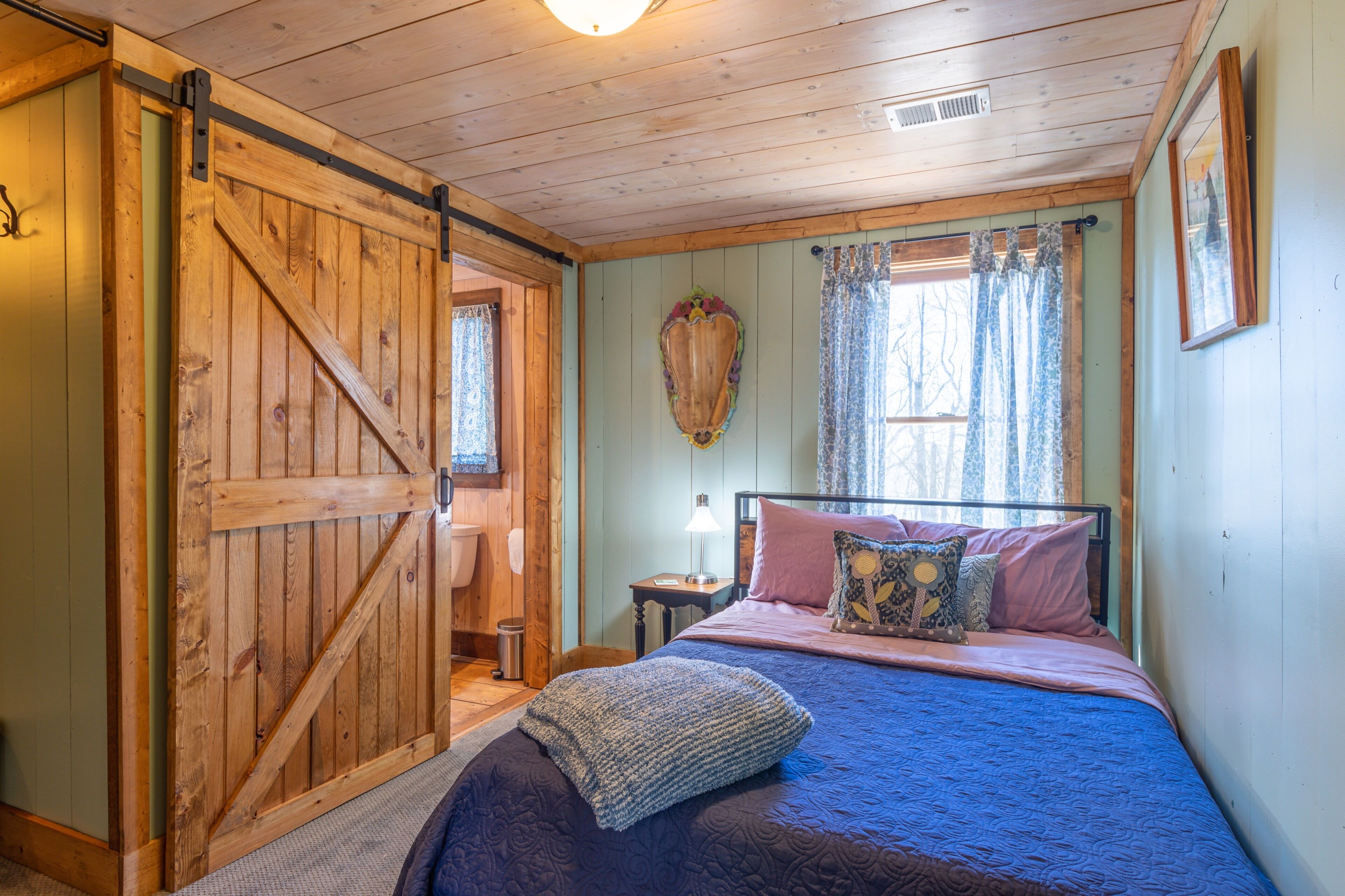 A bedroom with a bed, pillows, a nightstand, a lamp, a wooden door, and a window with curtains.