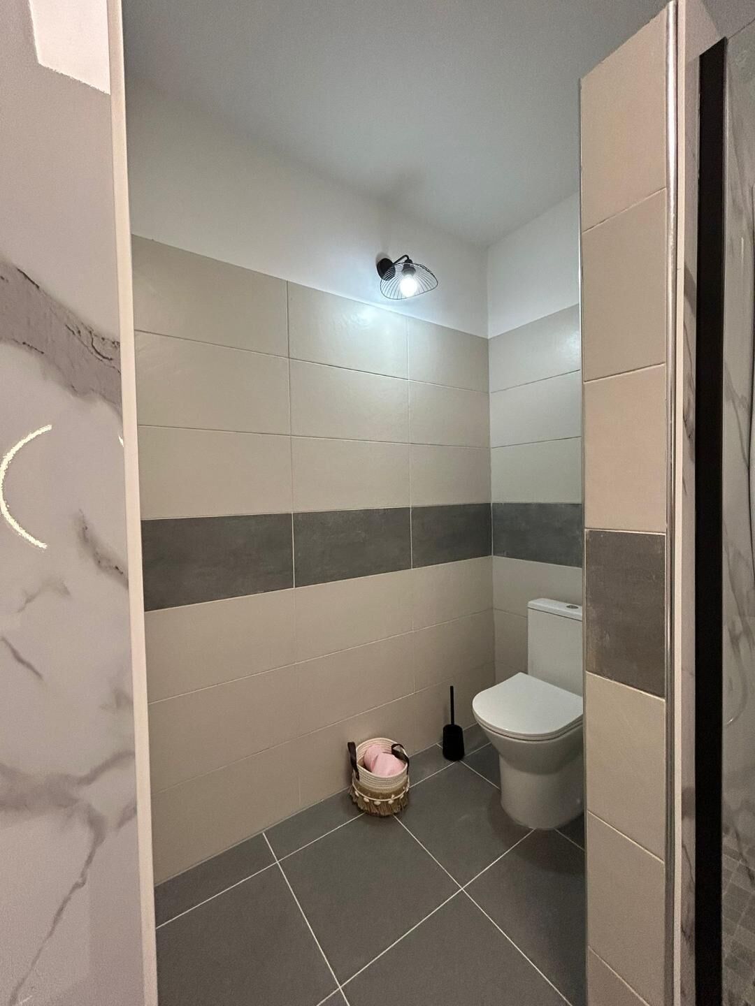 Un bagno con piastrelle grigie e bianche, un water, un piccolo cesto e un portalampada a parete.