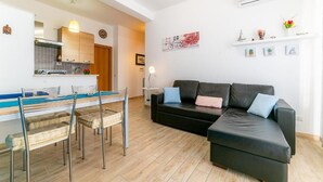 Un salón moderno con un sofá de cuero negro, una mesa de comedor con sillas, una zona de cocina con armarios de madera y un aire acondicionado instalado en la pared.