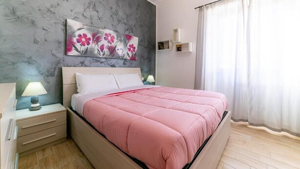 Un dormitorio con cama, mesitas de noche, un cuadro colgado en la pared y una ventana con cortinas.