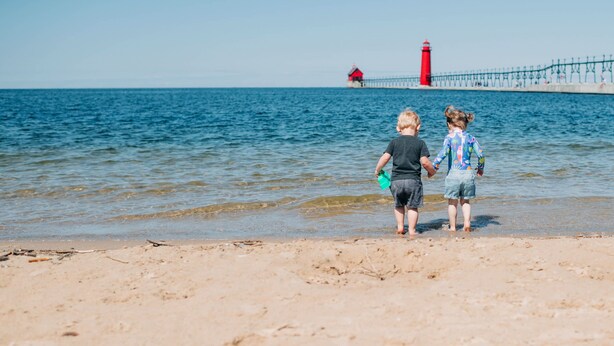 Two people walking on a sandy beach with a lighthouse in the background. - Harborside Grand Haven - Walk to Lake Michigan (Grand Haven)