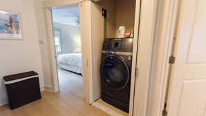A laundry room with a washing machine, a basket, and a door leading to a bedroom. - Harborside Grand Haven - Walk to Lake Michigan (Grand Haven)