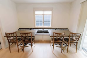 A dining area with a long table and four chairs, a window with blinds, and a bench with cushions.