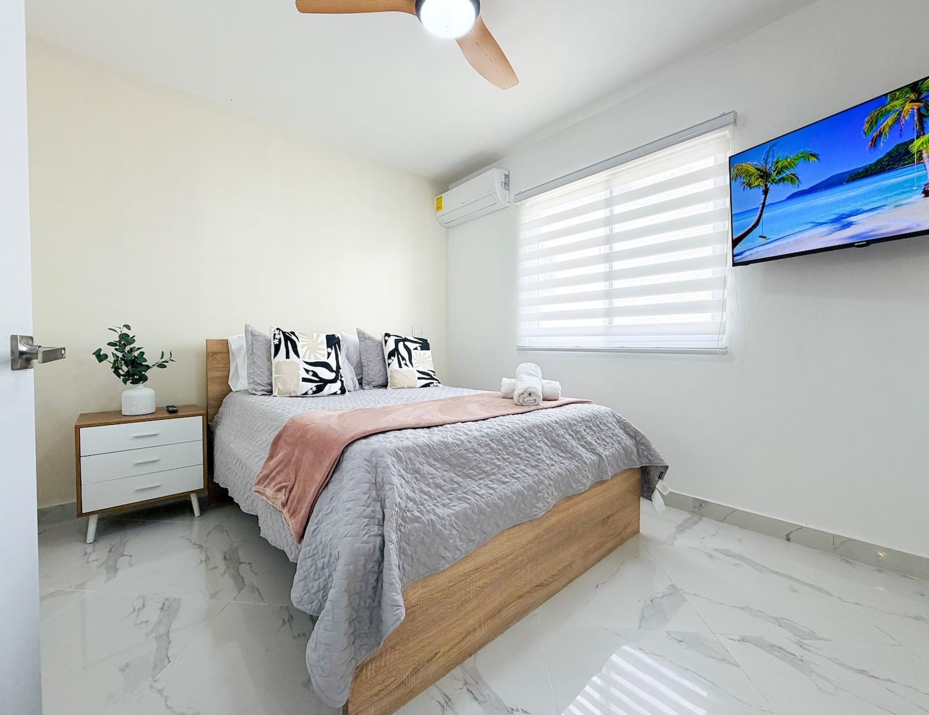 Une chambre à coucher comprenant un lit, une table de chevet, un ventilateur de plafond, un téléviseur et une fenêtre avec des stores.