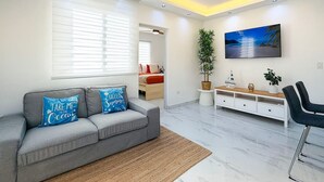 Een moderne woonkamer met een grijze bank, een flatscreen televisie en een groot raam met zonwering.
