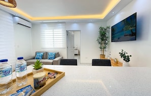 A modern living room with a sofa, a coffee table, and a dining area with a tray of snacks and water bottles.