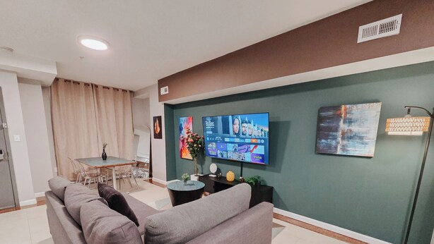 A modern living room with a grey sofa, a dining area with a table and chairs, a flat-screen TV mounted on the wall, and a floor lamp. - Element / King Bed / Elevator / Adjustable Desk (Philadelphia)