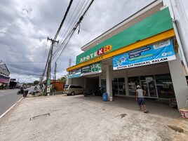 Una farmacia con toldo verde y amarillo, un cartel anunciando el Día Mundial del Farmacéutico 2025, y un hombre de pie en la acera.