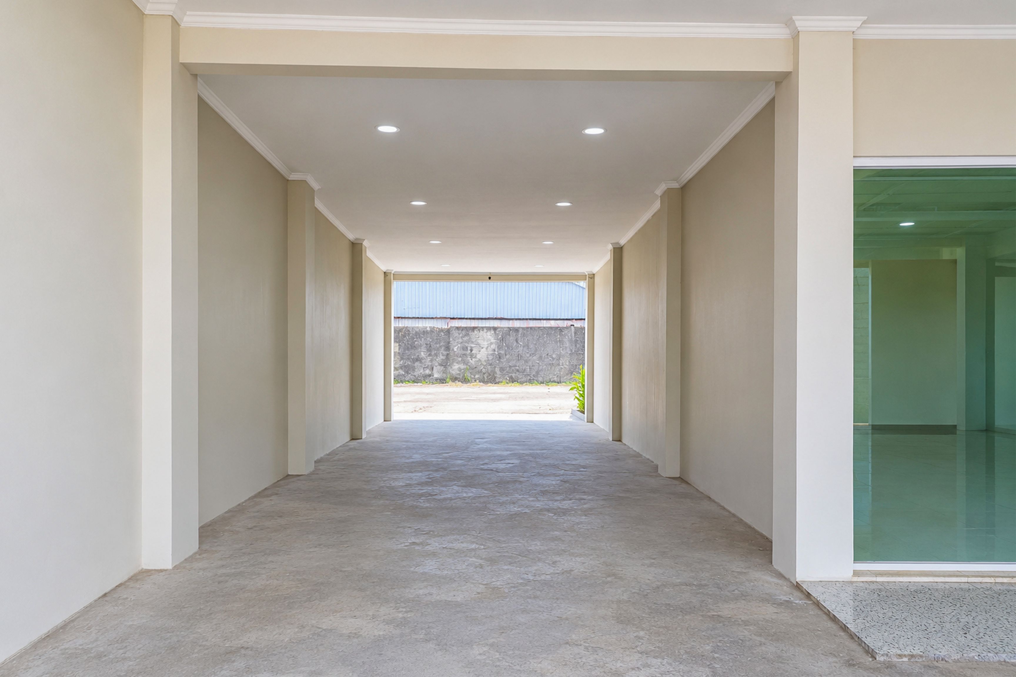 A corridor with a concrete floor leading to an outdoor area with a view of a stone wall and a blue structure.