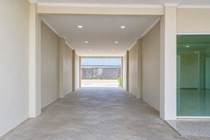 Un couloir doté d’un sol en béton mène à un espace extérieur d’où l’on peut admirer un mur de pierre et une structure bleue.