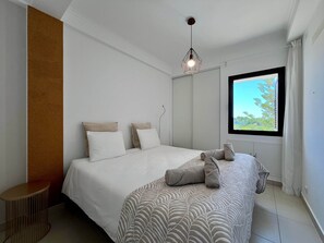 A bedroom with a bed, pillows, a hanging light fixture, a window with a view, and a small side table. - "Mar à Vista" Apartment (Olhos de Água)