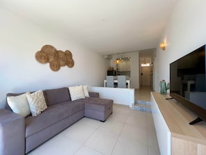 A modern living room with a grey sectional sofa, a flat-screen TV, and a kitchen in the background. - "Mar à Vista" Apartment (Olhos de Água)