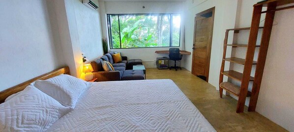 A bedroom with a bed, a desk, a chair, a sofa, and a window with a view of greenery.
