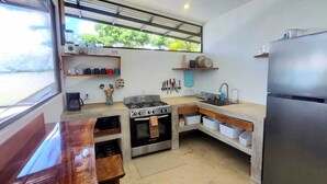Een kleine keuken met een kookplaat, spoelbak en koelkast, houten aanrechtbladen en een raam met uitzicht.