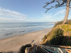 A beach with a wooden staircase leading down to the shore, surrounded by greenery and a clear sky. - Coastal Escape w/ Beach Access by Lighthouse (Coos Bay)