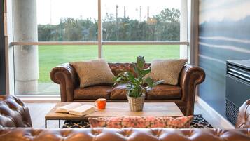 Uma sala de estar acolhedora com um sofá de couro, uma mesa de centro com um livro e uma caneca, e vista para um gramado verde através de grandes janelas.