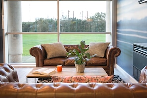A cozy living room with a leather sofa, a coffee table with a book and mug, and a view of a green lawn through large windows. - Student Only Maritime Studios (Falmouth)