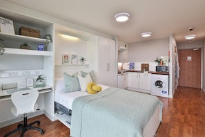 A compact bedroom with a bed, bedside table, chair, and built-in wardrobe. - Student Only Maritime Studios (Falmouth)