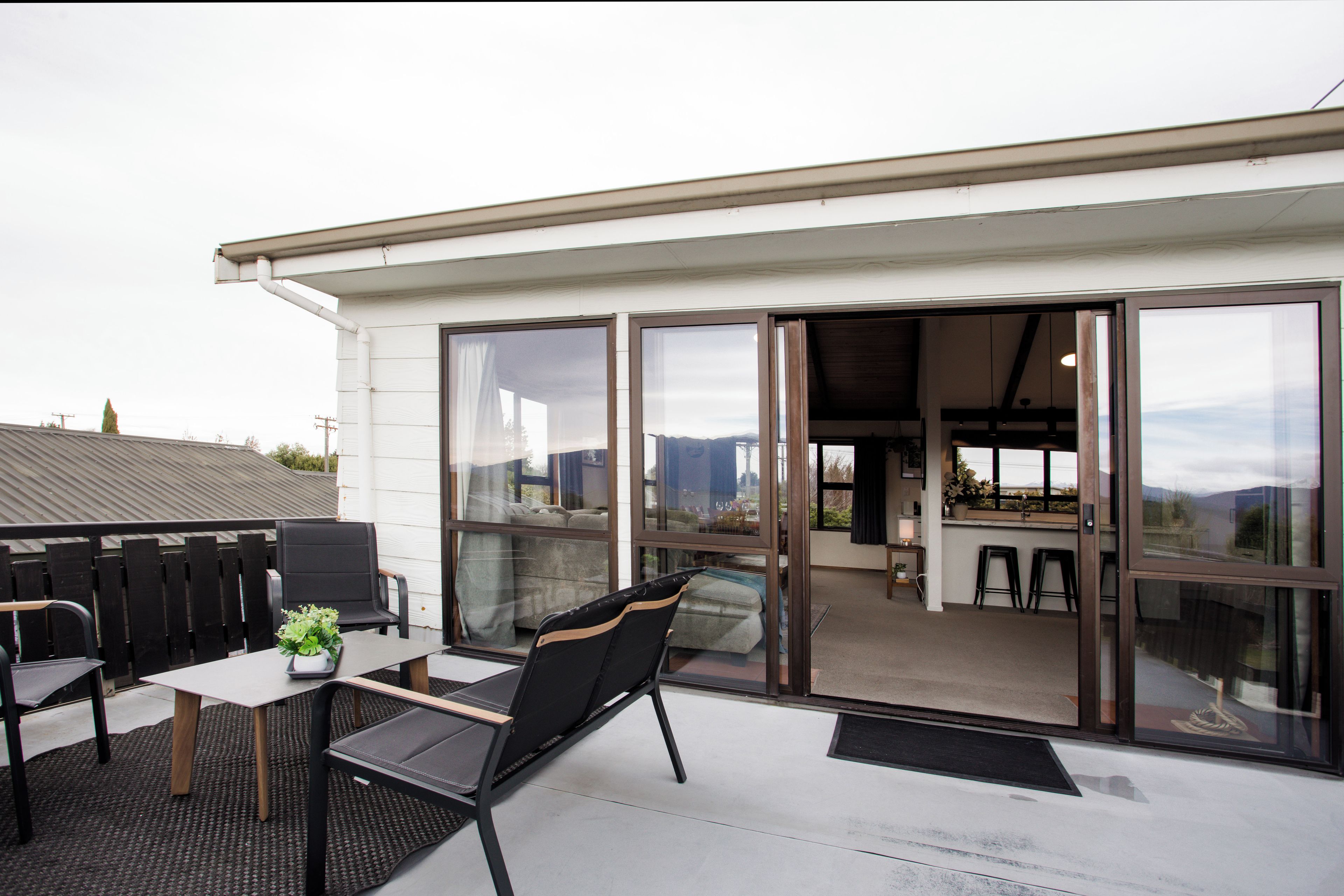 Een terras buiten met een tafel, stoelen, een plant in pot en uitzicht op het interieur door glazen deuren.