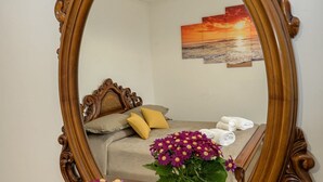 Een slaapkamer met een bed, kussens, een nachtkastje met handdoeken, een spiegel en een schilderij aan de muur.
