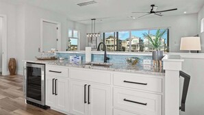 Une cuisine moderne avec des meubles blancs, un four encastré et une grande fenêtre donnant sur les maisons voisines.