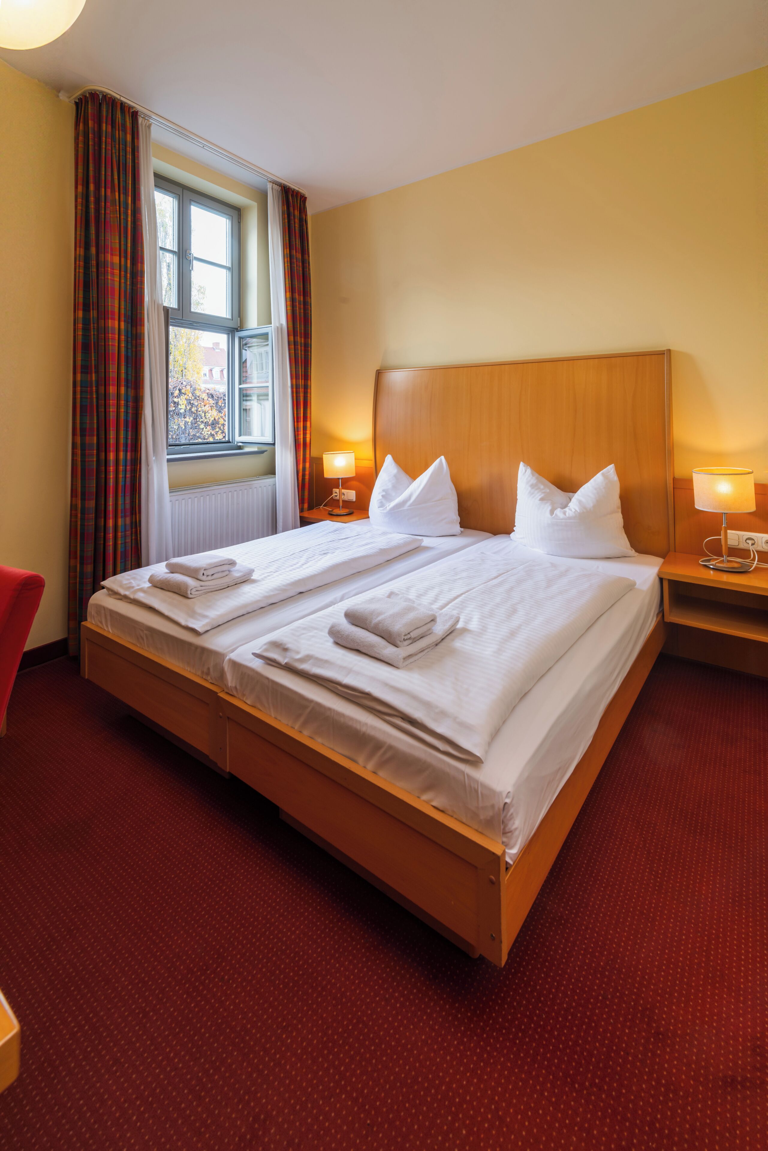Habitación de hotel con una cama grande, dos mesitas de noche con lámparas, una ventana con cortinas y una alfombra roja.