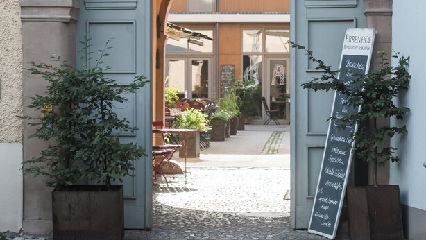 An entrance to a building labeled 'ERBENHOF' with a number '10' above the door. There is a signboard with text, and potted plants are placed on either side of the entrance. - Erbenhof (Weimar)