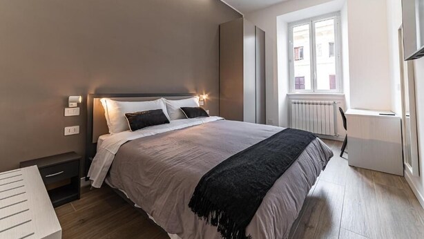 A bedroom with a bed, bedside table, lamp, window, radiator, and a view of buildings outside. - Vatican Suites Apartment - Vatican City (Rome)