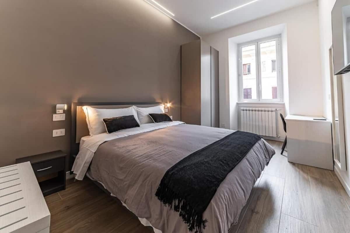 A bedroom with a bed, bedside table, lamp, window, radiator, and a view of buildings outside.