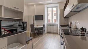 A modern kitchen with a built-in microwave, a dining area with chairs, a TV mounted on the wall, and a window providing natural light. - Vatican Suites Apartment - Vatican City (Rome)