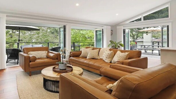 Een ruime woonkamer met een grote bank, een salontafel en een televisie. Er zijn grote ramen en een balkon met tuinmeubels.