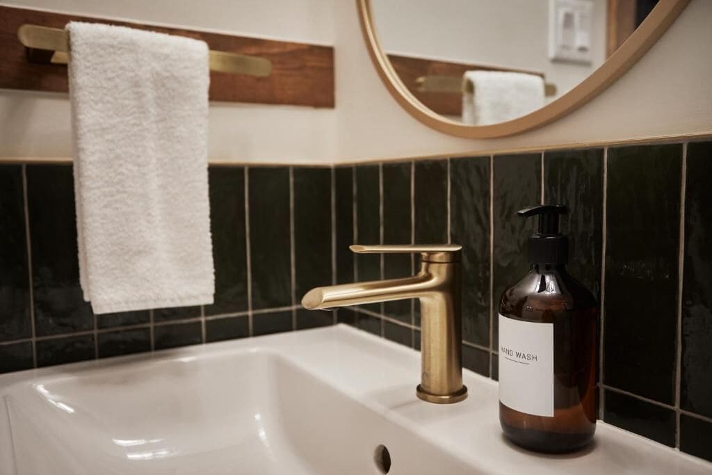 Ein Badezimmer mit einem weißen Waschbecken, einem goldenen Wasserhahn, einer Handseifenflasche, einem weißen Handtuch, das an einem Handtuchhalter hängt, und einem Spiegel mit goldener Rahmung.