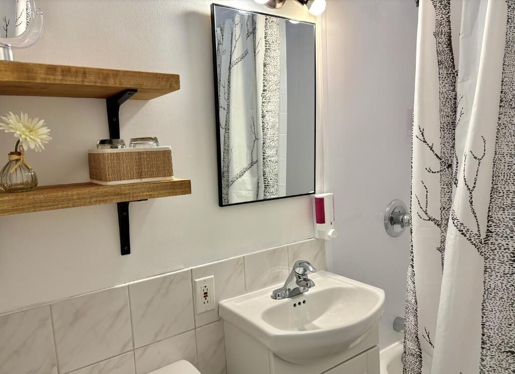 Un bagno con lavandino bianco, uno specchio, uno scaffale con un vaso e contenitori, e una tenda da doccia con motivo alberi.