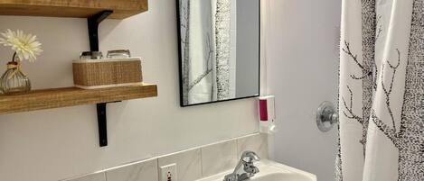 Un bagno con lavandino bianco, uno specchio, uno scaffale con un vaso e contenitori, e una tenda da doccia con motivo alberi.