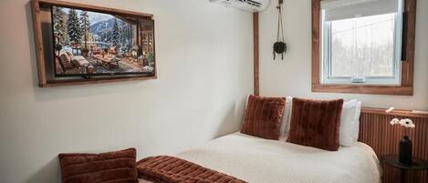 A bedroom with a bed, a mounted TV, an air conditioning unit, a window, and a small table with a vase.