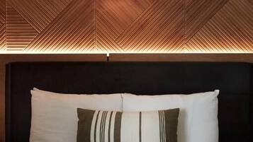 Uma cama com almofadas brancas e listradas, cabeceira escura e teto de madeira com padrão geométrico.