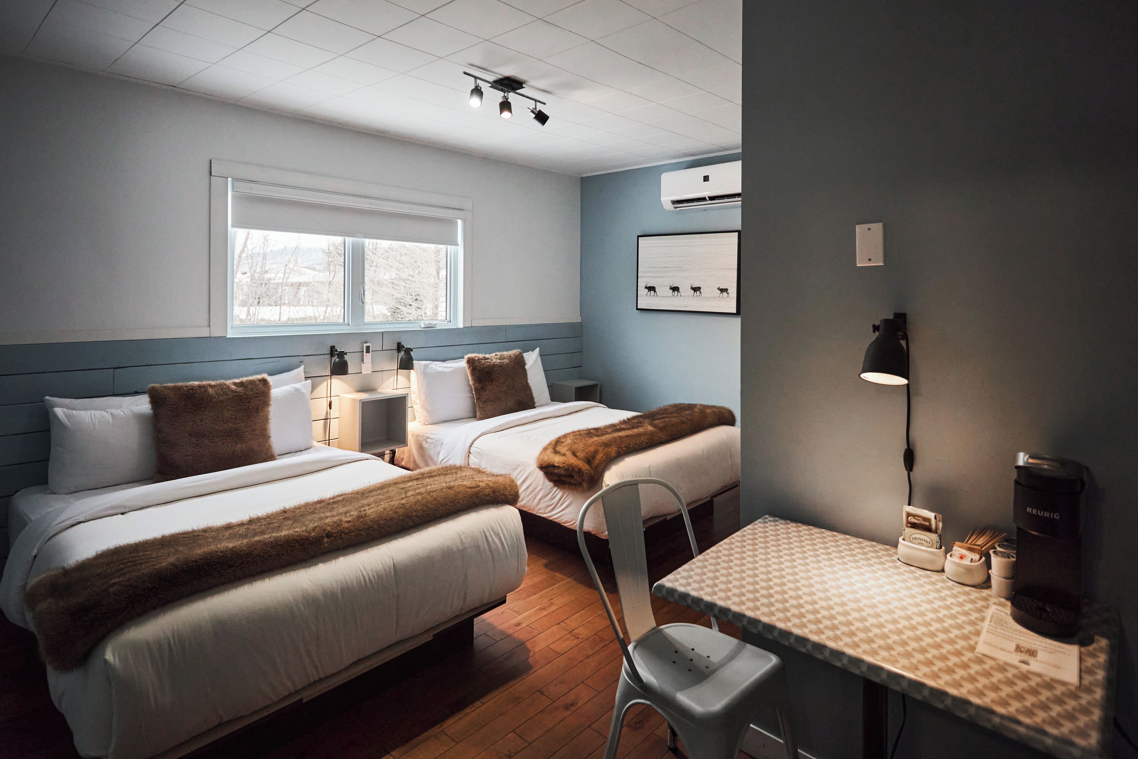 A room with two beds, a table with a lamp, a chair, an air conditioning unit, and a window with blinds.