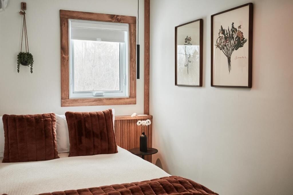 Une chambre avec une grande fenêtre, un lit avec une literie marron, deux œuvres d’art florales encadrées et une petite table de chevet sur laquelle se trouve un vase.