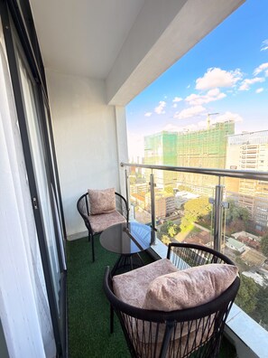 Een balkon met een glazen deur, twee rieten stoelen met kussens, een klein rond tafeltje en uitzicht op een stadsbeeld met gebouwen en een bouwkraan.