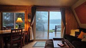 Een gezellige kamer met houten inrichting, een eettafel met stoelen, een bank met kussens, een bijzettafeltje met een boek en mok, en een groot raam met uitzicht op een besneeerd landschap.