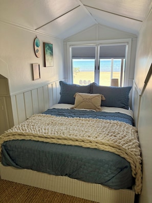A bedroom with a view of the beach, a bed with blue and beige bedding, and a window with blinds.