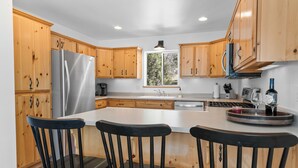 A kitchen with wooden cabinets, a stainless steel refrigerator, a large island with bar stools, and a window with a view of trees. - Paradise Pines (Big Bear Lake)