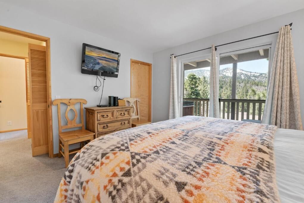 Un dormitorio con cama, televisor, cómoda, silla y balcón con cortinas.