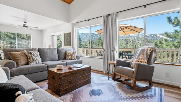 A spacious living room with a sofa, armchair, and coffee table, large windows with a mountain view, and a balcony with an umbrella. - Paradise Pines (Big Bear Lake)