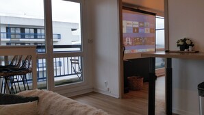 A modern living room with a large window, a sofa, a dining area with chairs, and a view of a building outside. - Panoramik station +parking (Nancy)