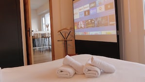 A hotel room with a bed, a projector screen displaying streaming services, a wooden chair, and a dining area in the background. - Panoramik station +parking (Nancy)