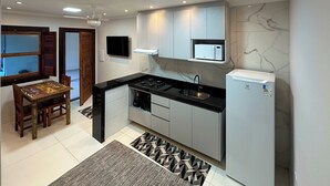 Eine kompakte Küche mit Gasherd, Mikrowelle und Kühlschrank. Ein Esstisch mit Stühlen und einem gemusterten Teppich sind ebenfalls vorhanden.